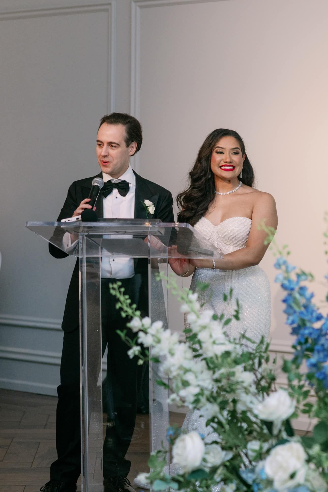 Bride and groom smile together at acrylic podium during wedding reception speeches with flowers.