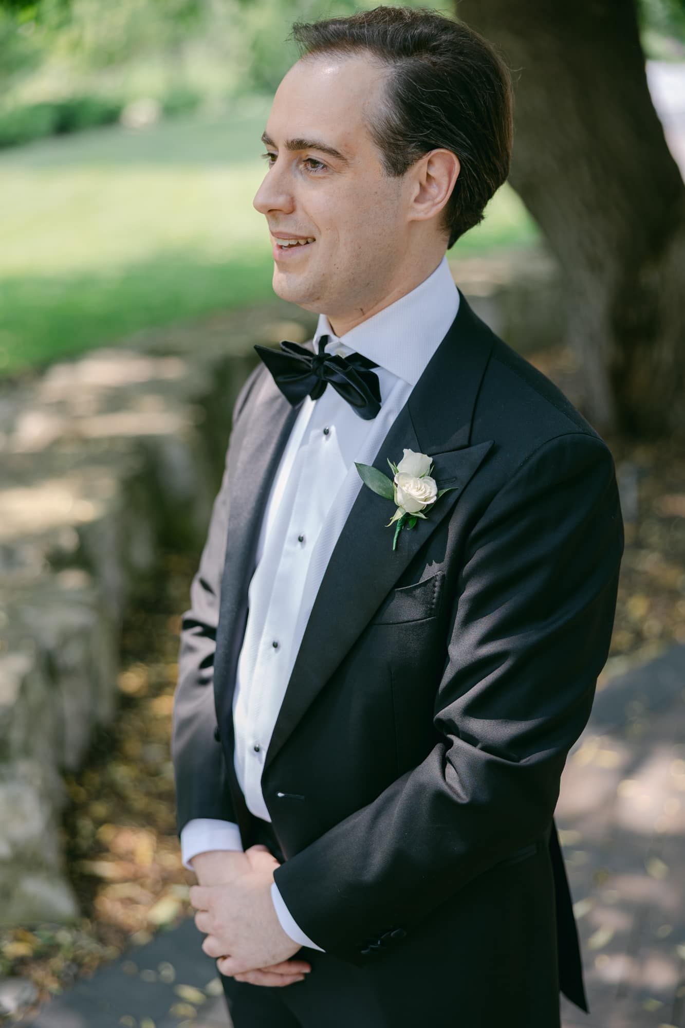 Dapper groom in black tuxedo with bow tie and white rose boutonniere smiles outdoors on wedding day.