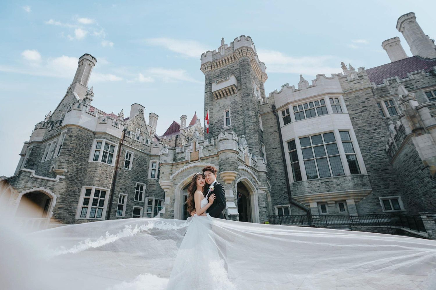 Bride and groom kissing with flowing veil billowing before magnificent Casa Loma castle facade.
