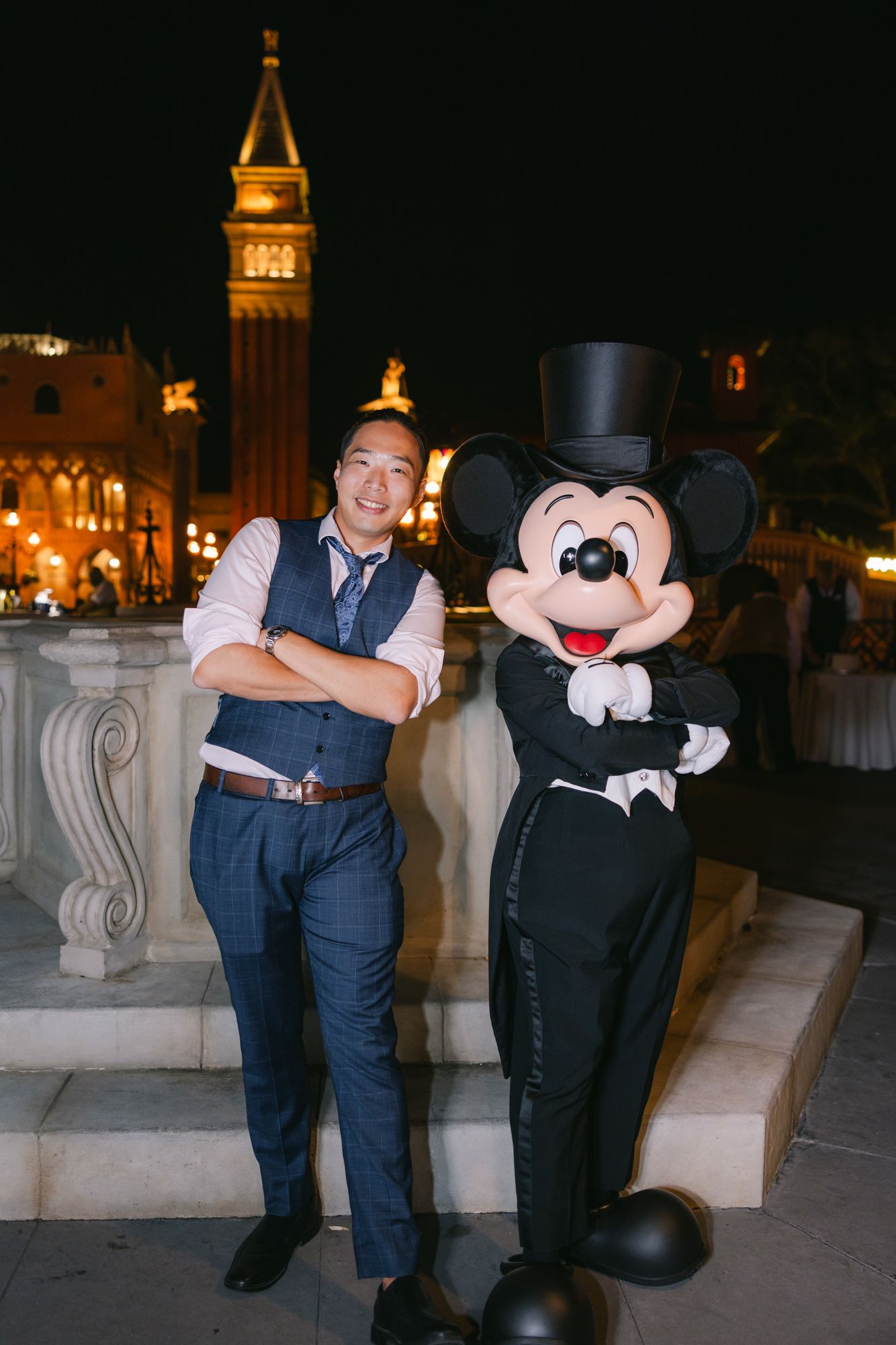 Groom in navy vest poses with Mickey Mouse in top hat and tuxedo at Disney park night event.