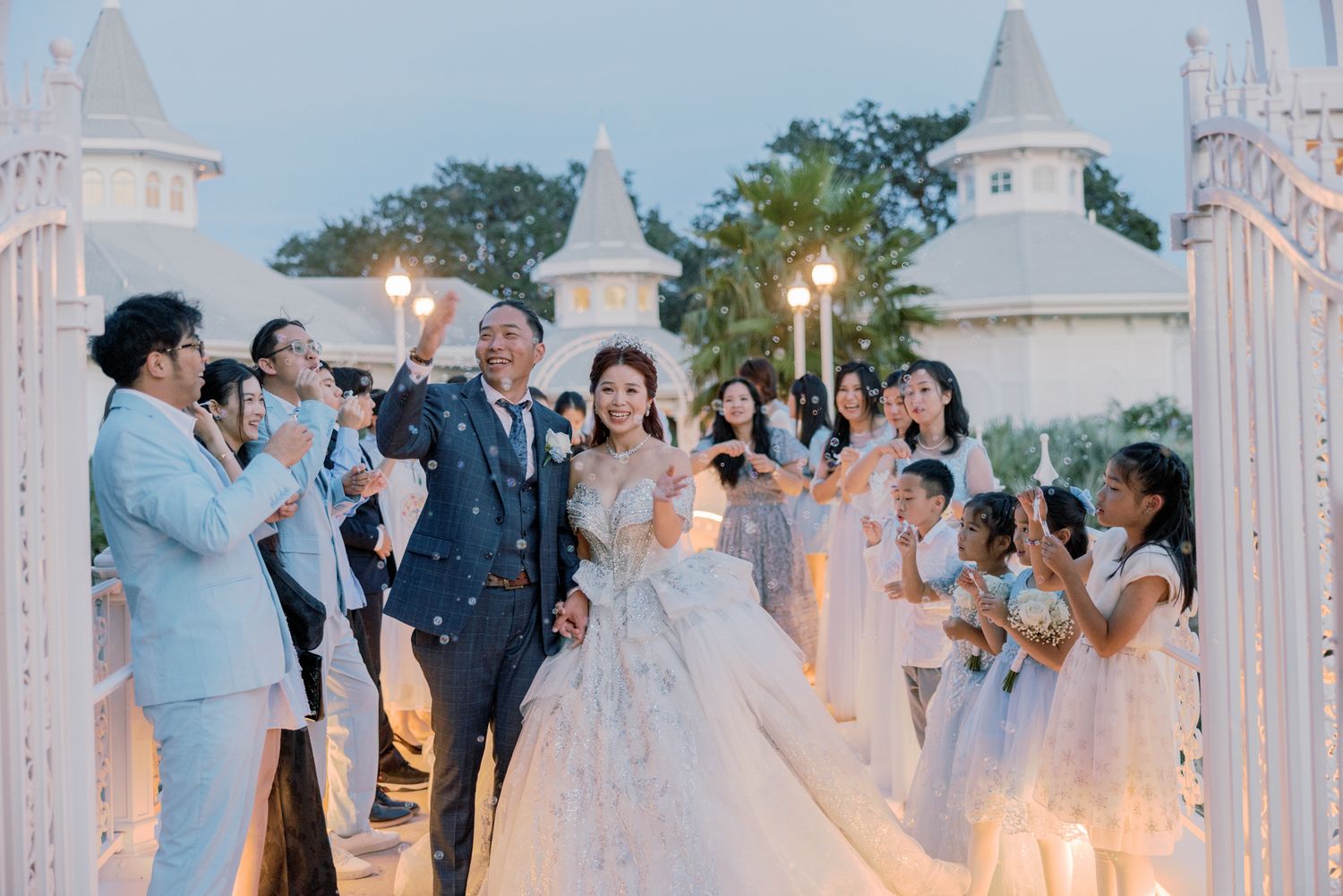 Bride and groom exit wedding ceremony through white gates as guests celebrate with confetti and champagne.