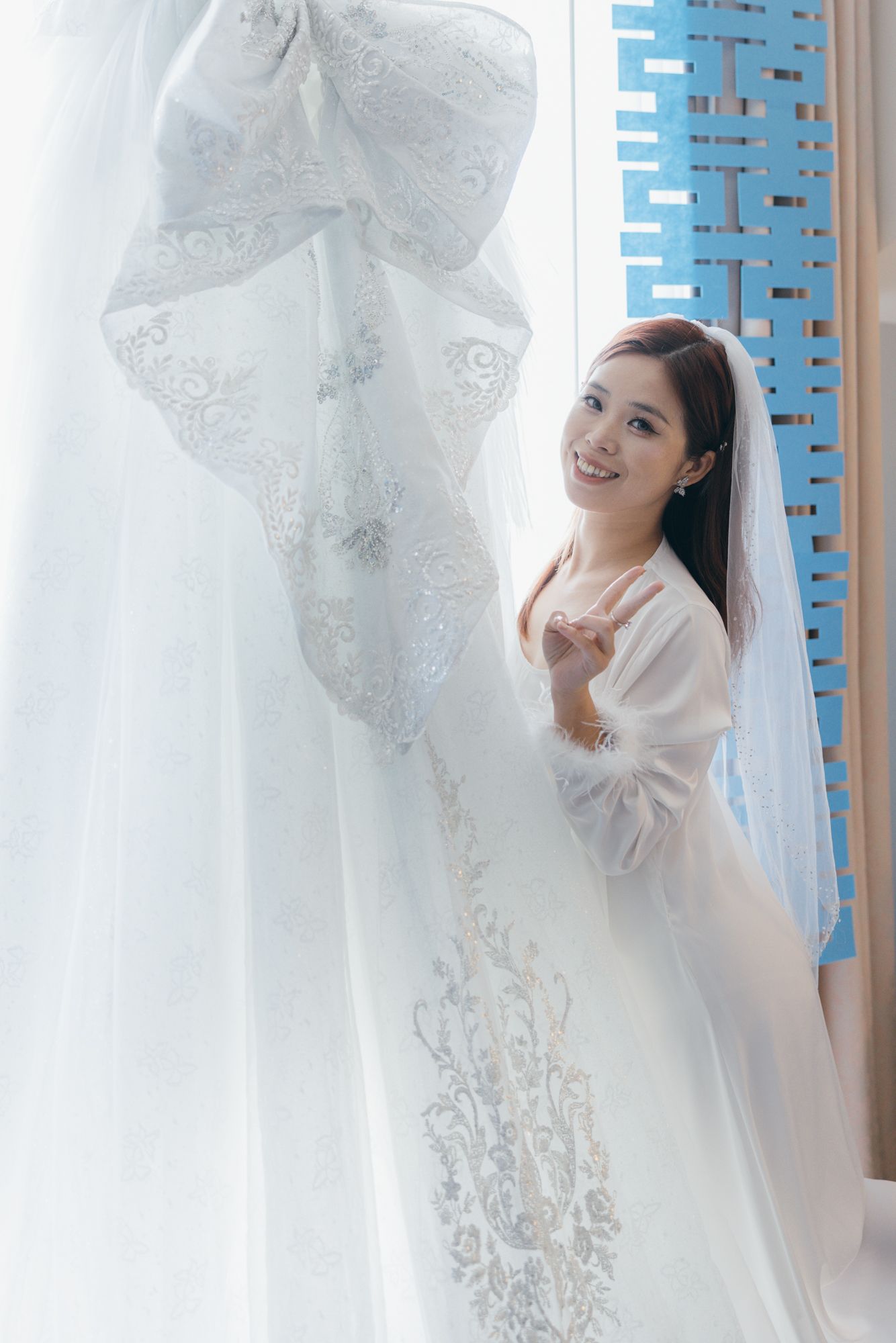 Smiling Asian bride in white wedding dress and veil posing elegantly beside a window with lace curtains.