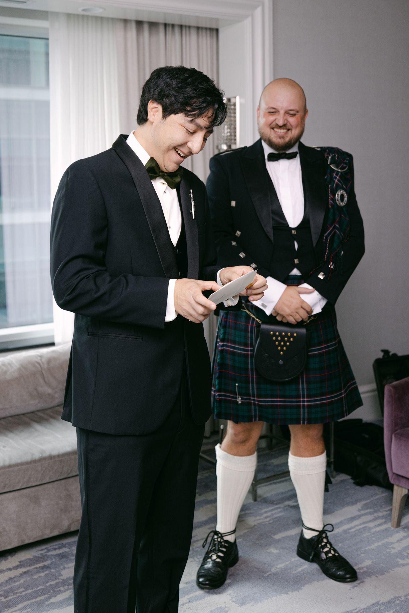 Groom in a black tuxedo and bow tie reads a card while a smiling groomsman in a tartan kilt and sporran stands beside him.