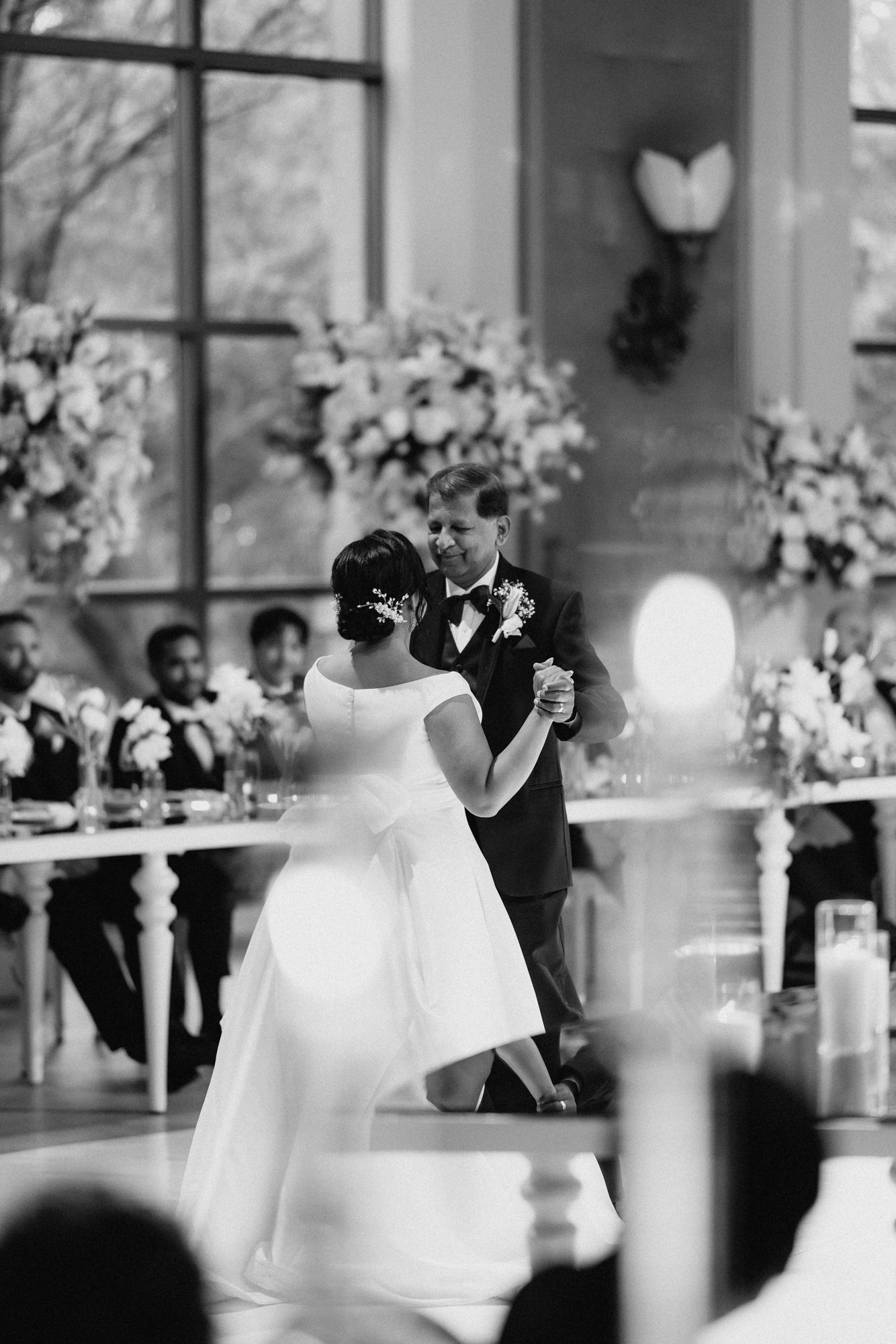 Black and white photo of a bride in an off-the-shoulder gown dancing with an older man in a tuxedo at an elegant wedding reception.