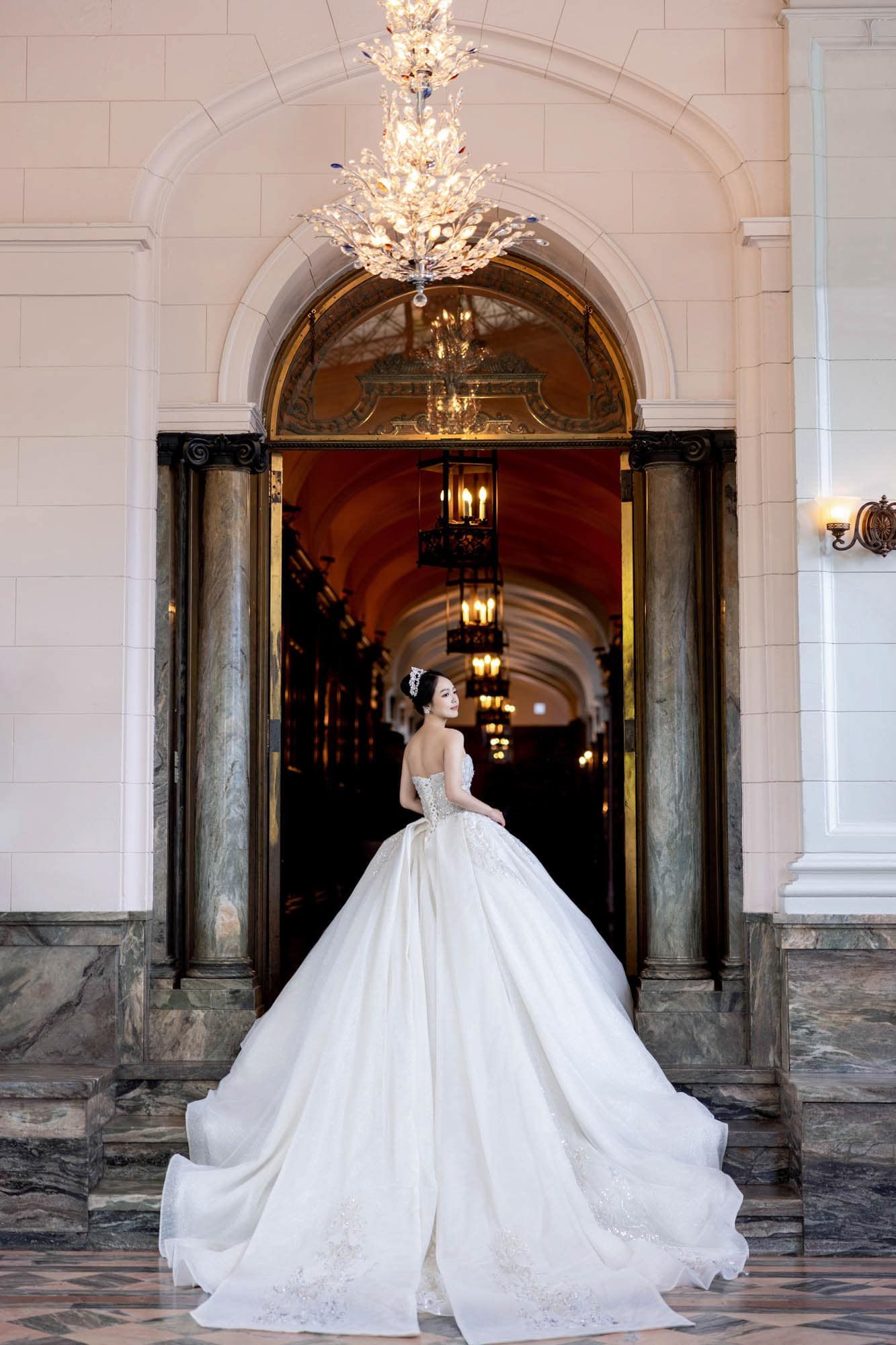 Bride in a stunning ballgown with long train standing in a grand arched doorway with chandeliers.
