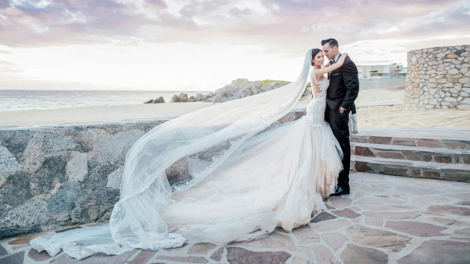 Bride and groom share a romantic kiss outdoors, with cathedral veil flowing in the wind near a coastal stone wall.