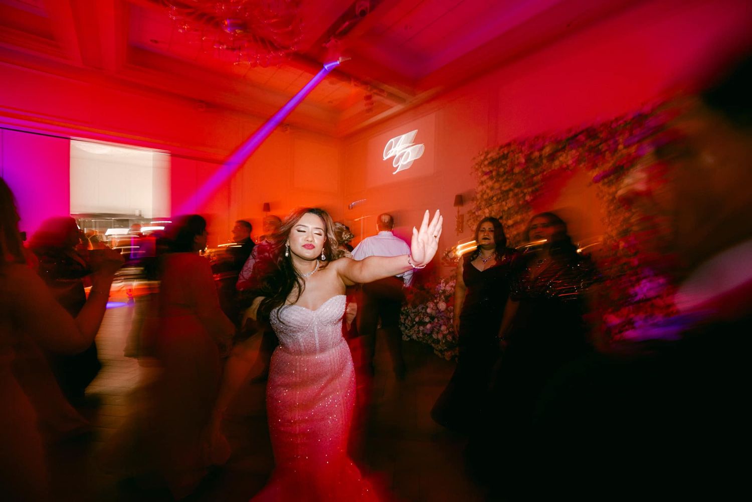 Bride in strapless gown dancing joyfully with guests under vibrant red lights at wedding reception.