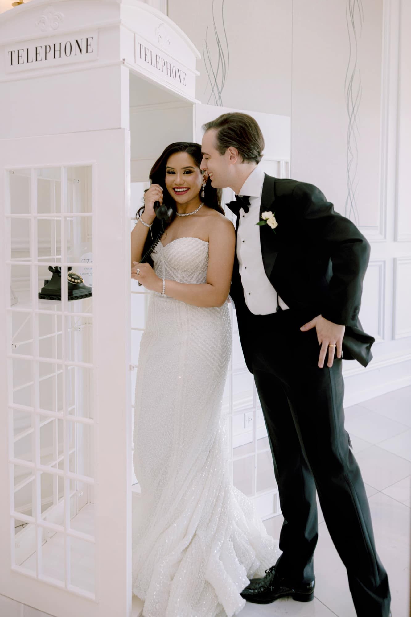 Bride and groom pose playfully beside white telephone booth at elegant wedding venue.