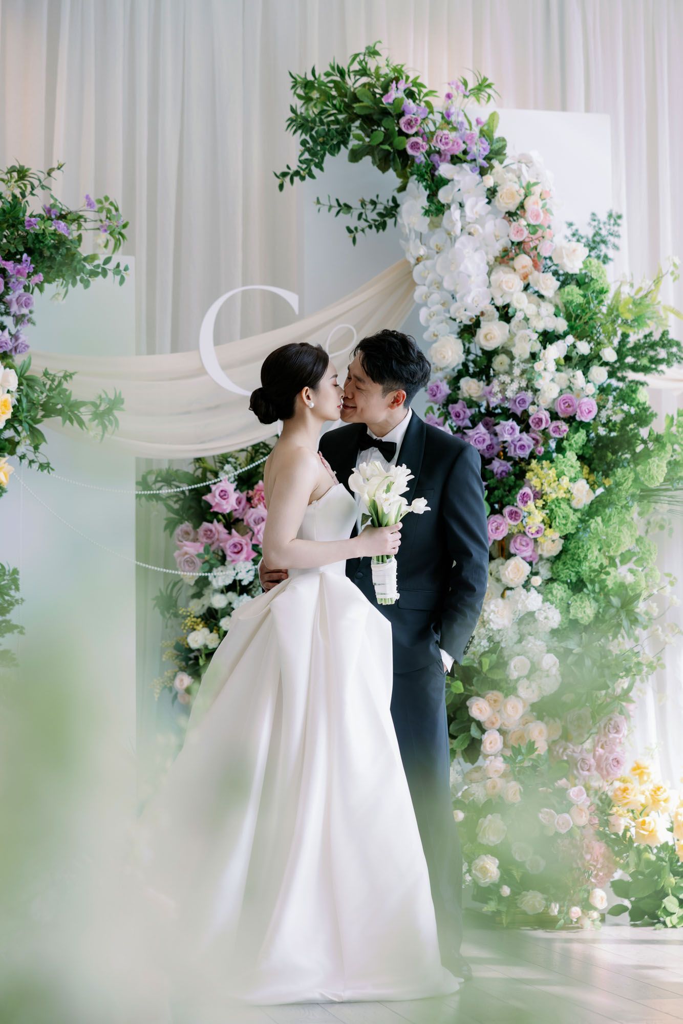 Bride and groom share a tender moment before a lush floral arch with purple and white blooms.
