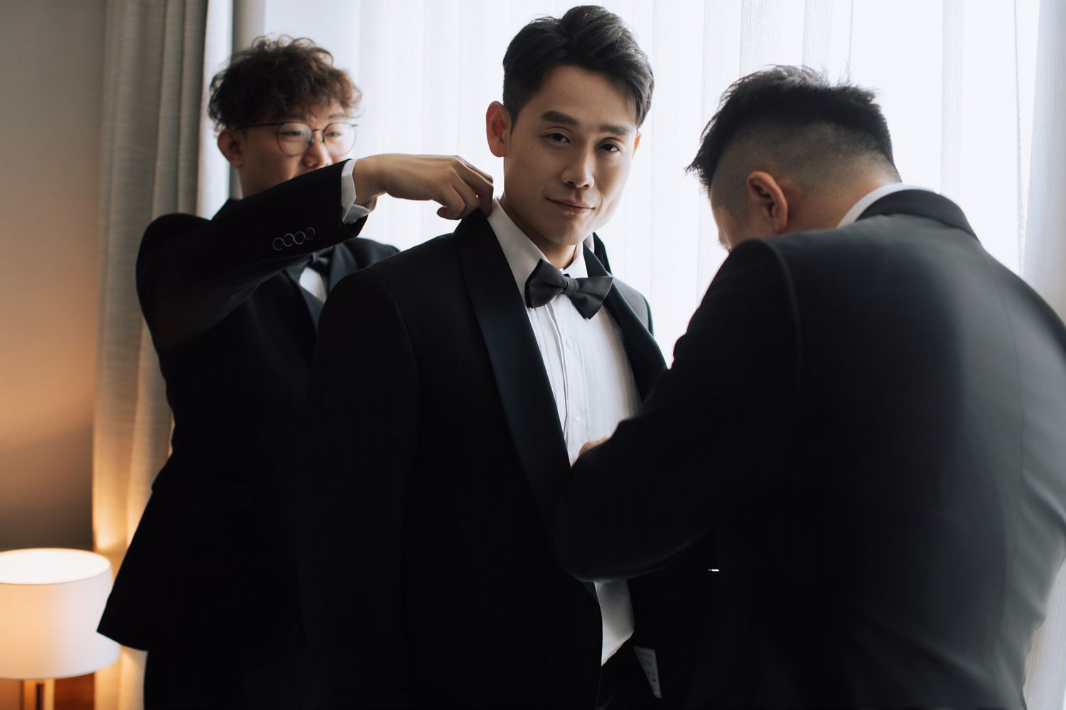 Groomsmen helping groom adjust bow tie and black tuxedo jacket in hotel room.