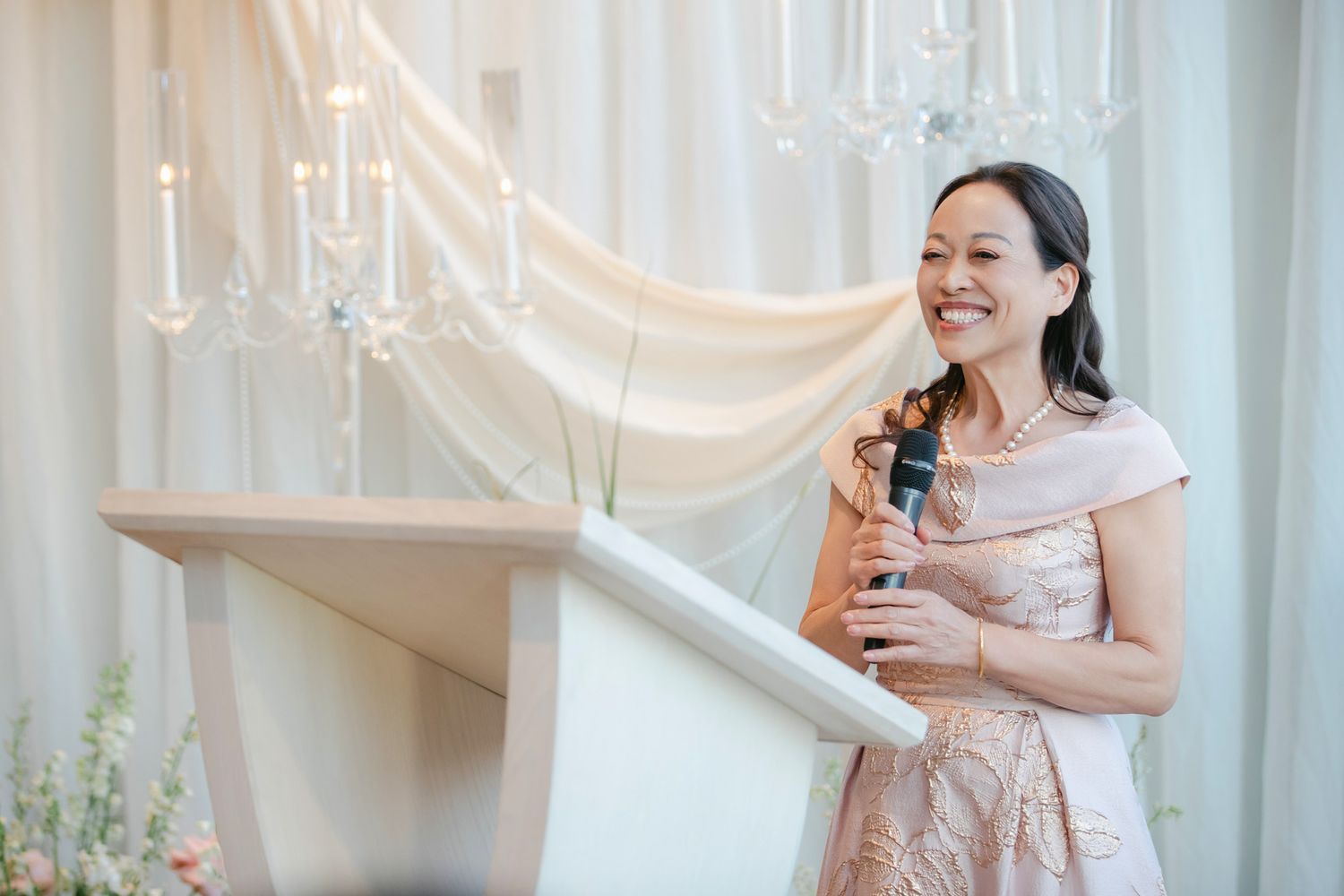 Woman in pink dress smiling and speaking into a microphone at a wedding reception podium.