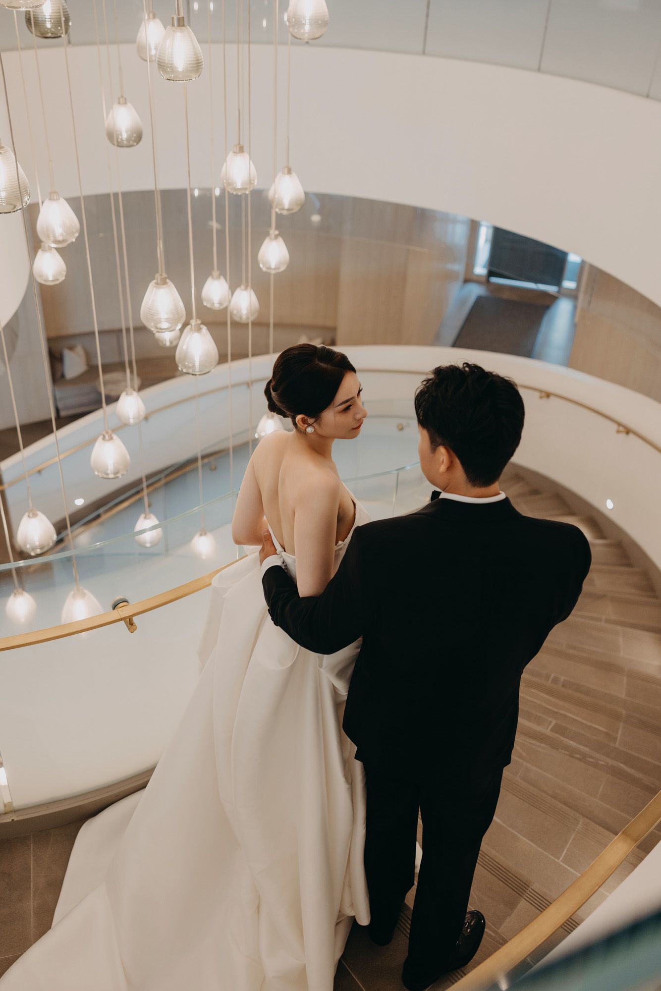 Bride and groom share an intimate moment on elegant spiral staircase with pendant lighting above.