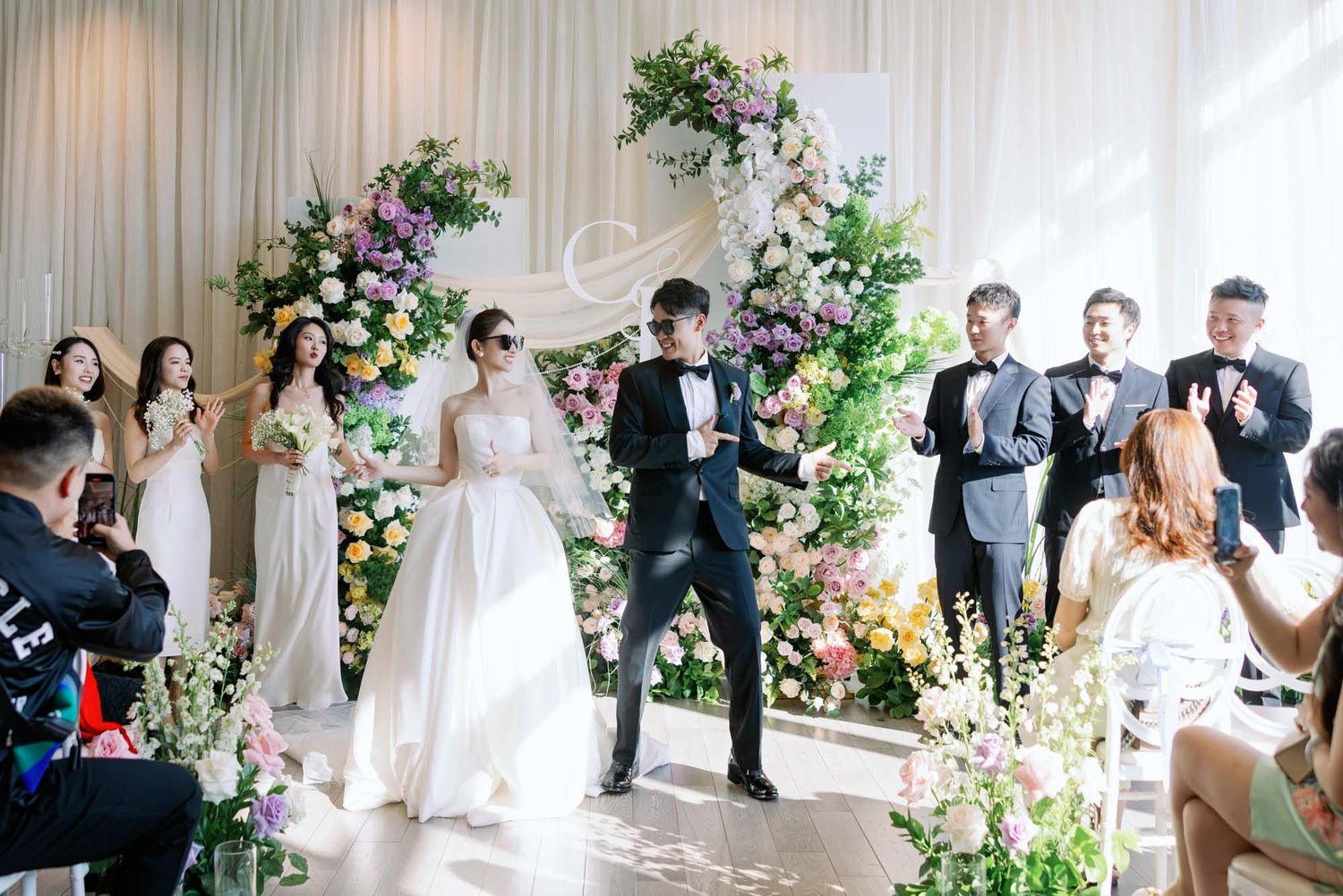 Bride and groom celebrate joyfully with wedding party before colorful floral arch at ceremony.