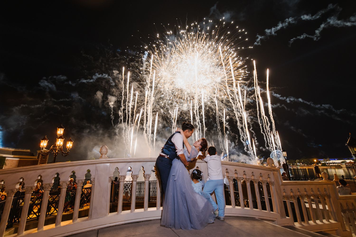 Newlyweds embrace on bridge with spectacular fireworks display lighting the night sky above.