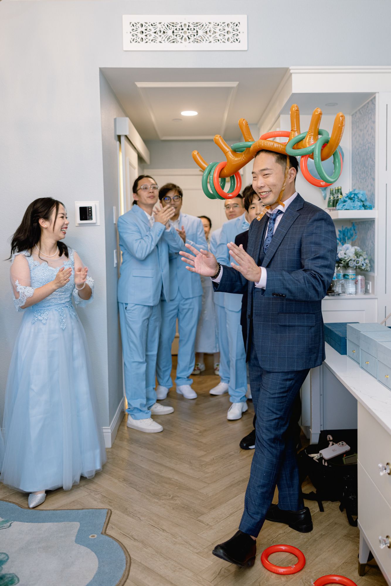 Groom wearing colorful ring toss antlers plays wedding game with smiling bridal party in blue.