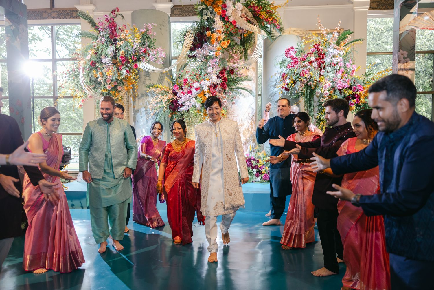 Groom in a cream sherwani walks down the aisle between cheering guests in colorful Indian attire beneath a vibrant floral arch.