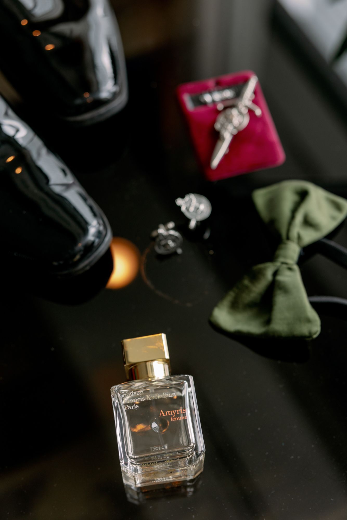 Groom's wedding accessories including a green bow tie, silver cufflinks, tie clip in a red box, black dress shoes and a perfume bottle.