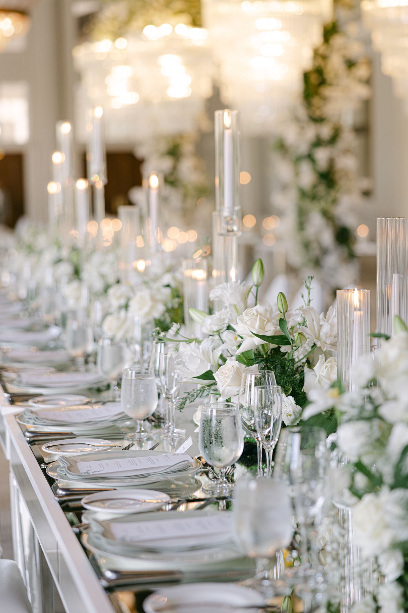 Elegant wedding reception table with white floral centerpieces, tall taper candles, glassware and place settings under chandeliers.