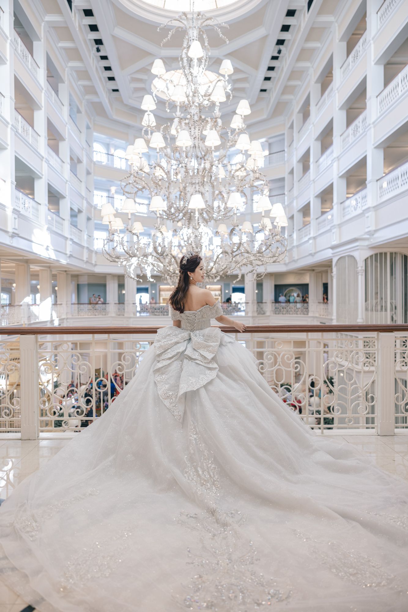 Bride in elegant white off-shoulder gown with dramatic train stands before grand crystal chandelier.