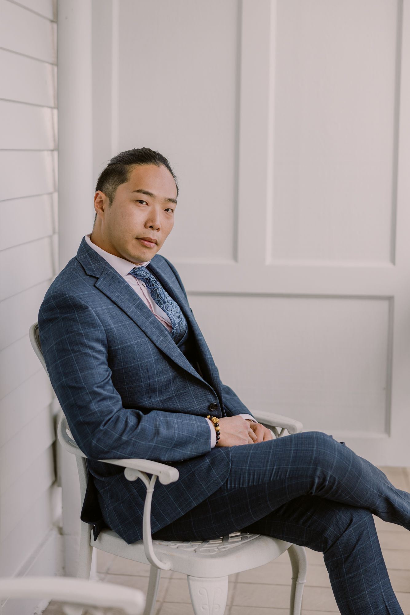 Young Asian man in blue plaid suit sitting confidently on white chair against light gray wall.