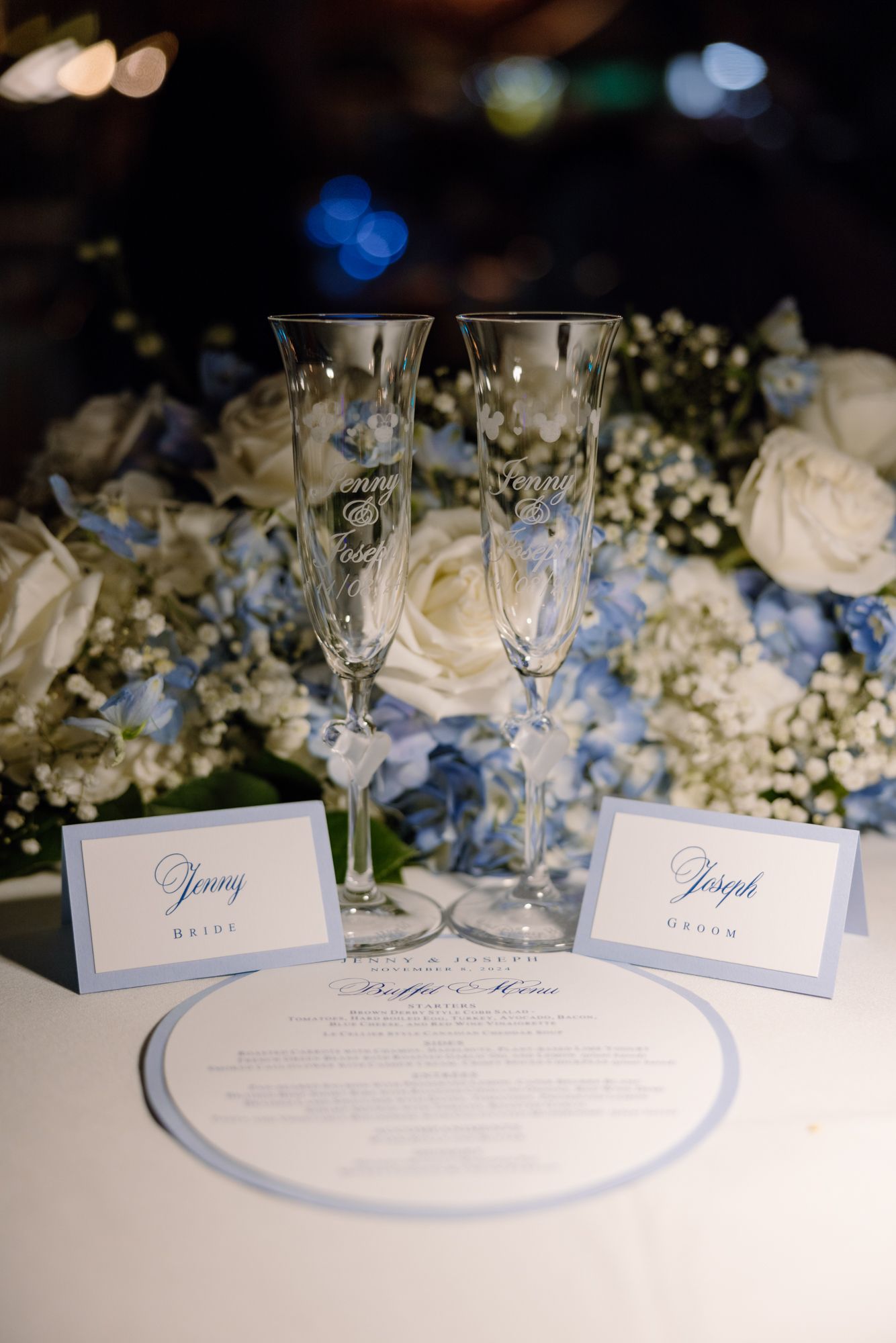 Personalized champagne flutes engraved with Jenny and Joseph beside place cards and blue floral centerpiece.