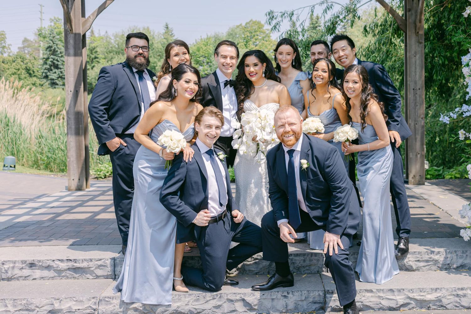 Wedding party poses together outdoors, bridesmaids in blue gowns with white bouquets and groomsmen in dark suits.