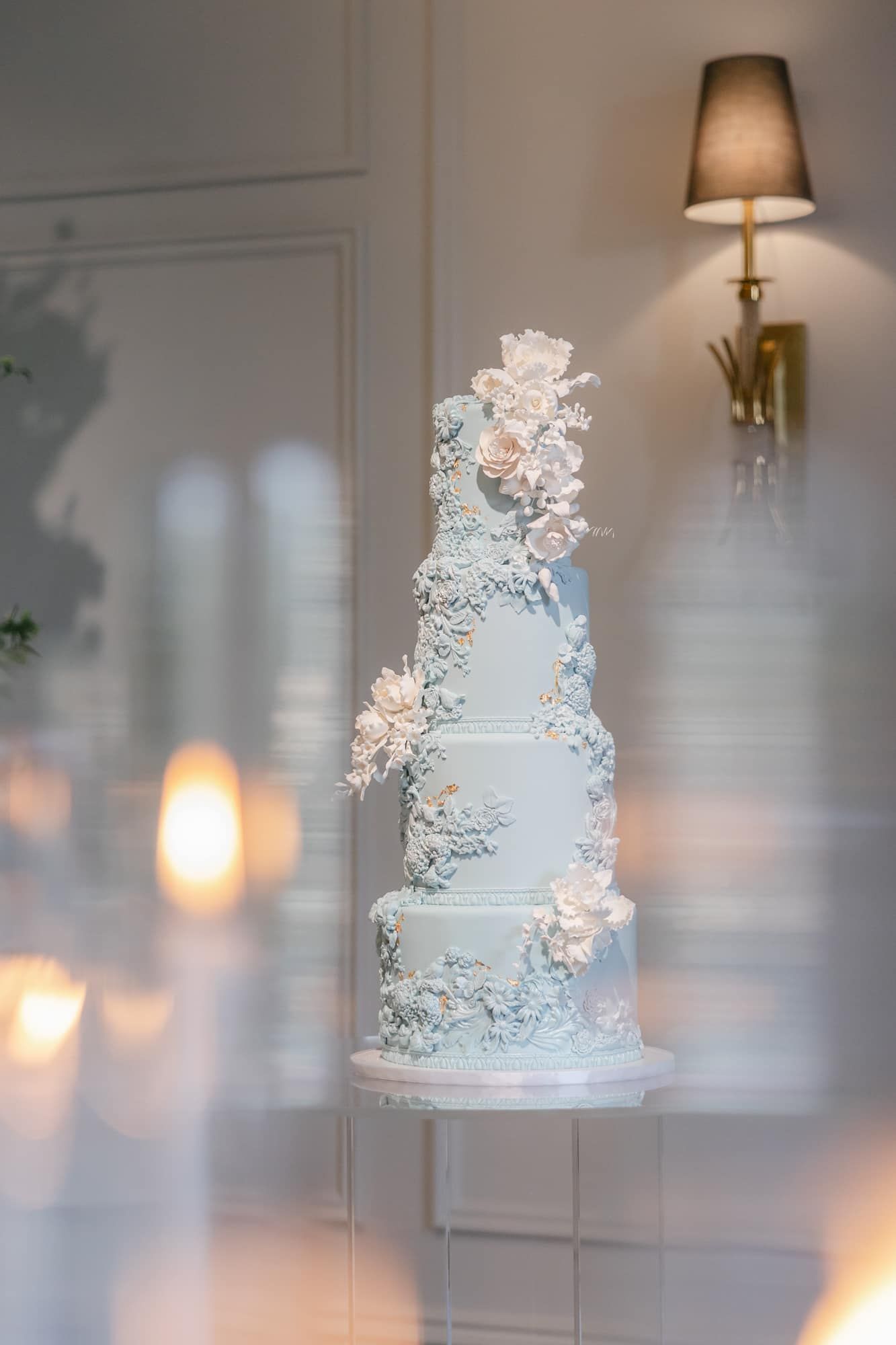 Five-tier light blue wedding cake adorned with white sugar flowers and intricate lace details on a pedestal.