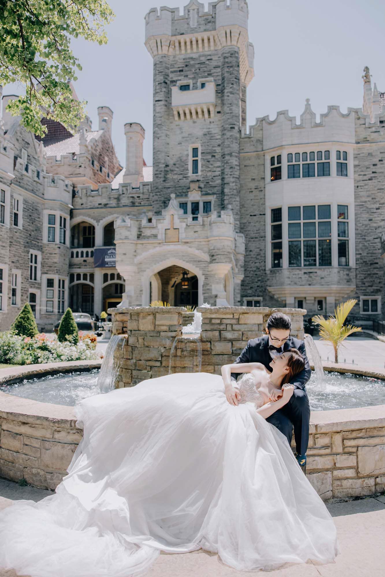 Groom dipping bride in a flowing white ballgown by a stone fountain in front of a grand castle.