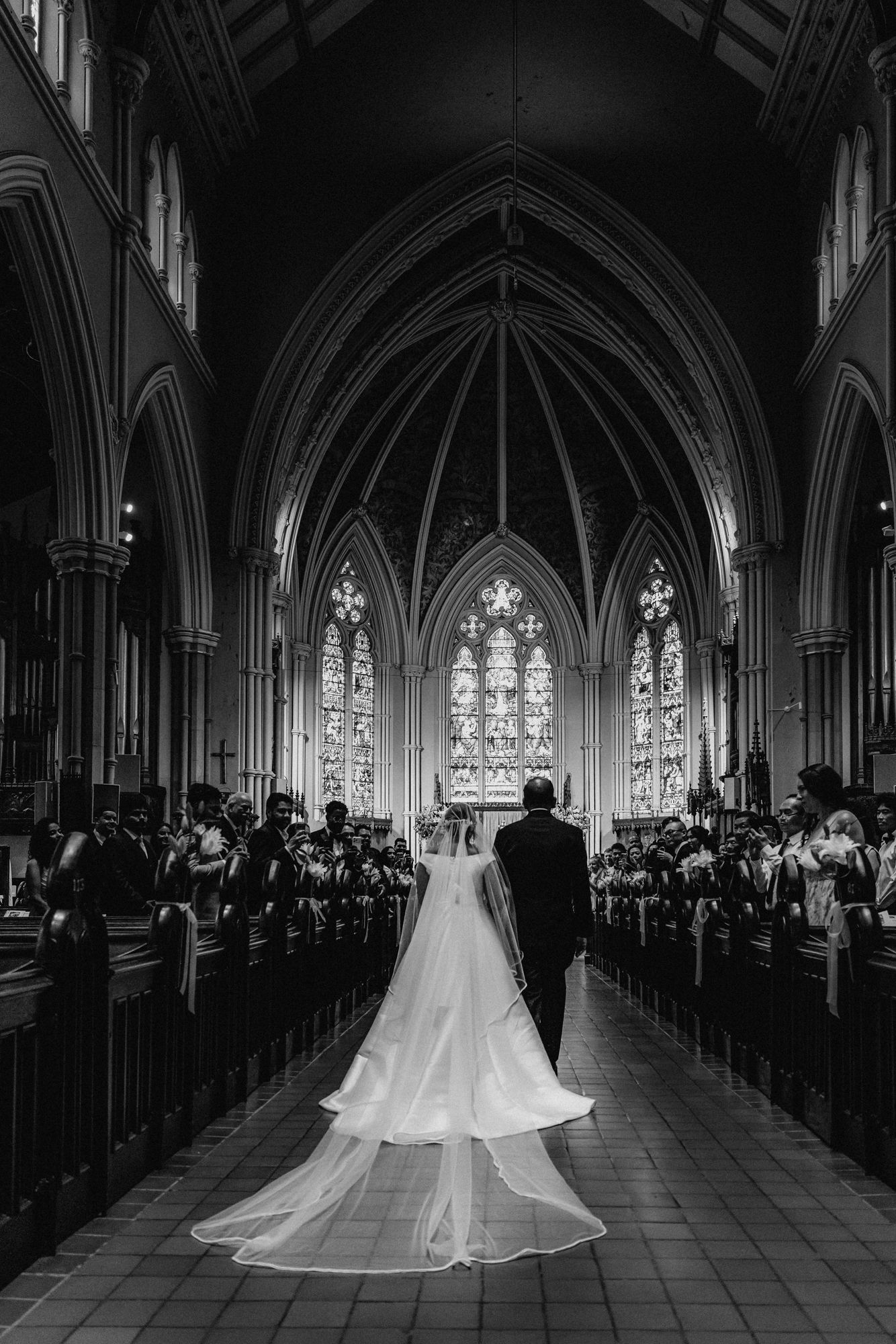 Bride in a long veil walks down the aisle of a grand Gothic church in black and white as wedding guests look on.