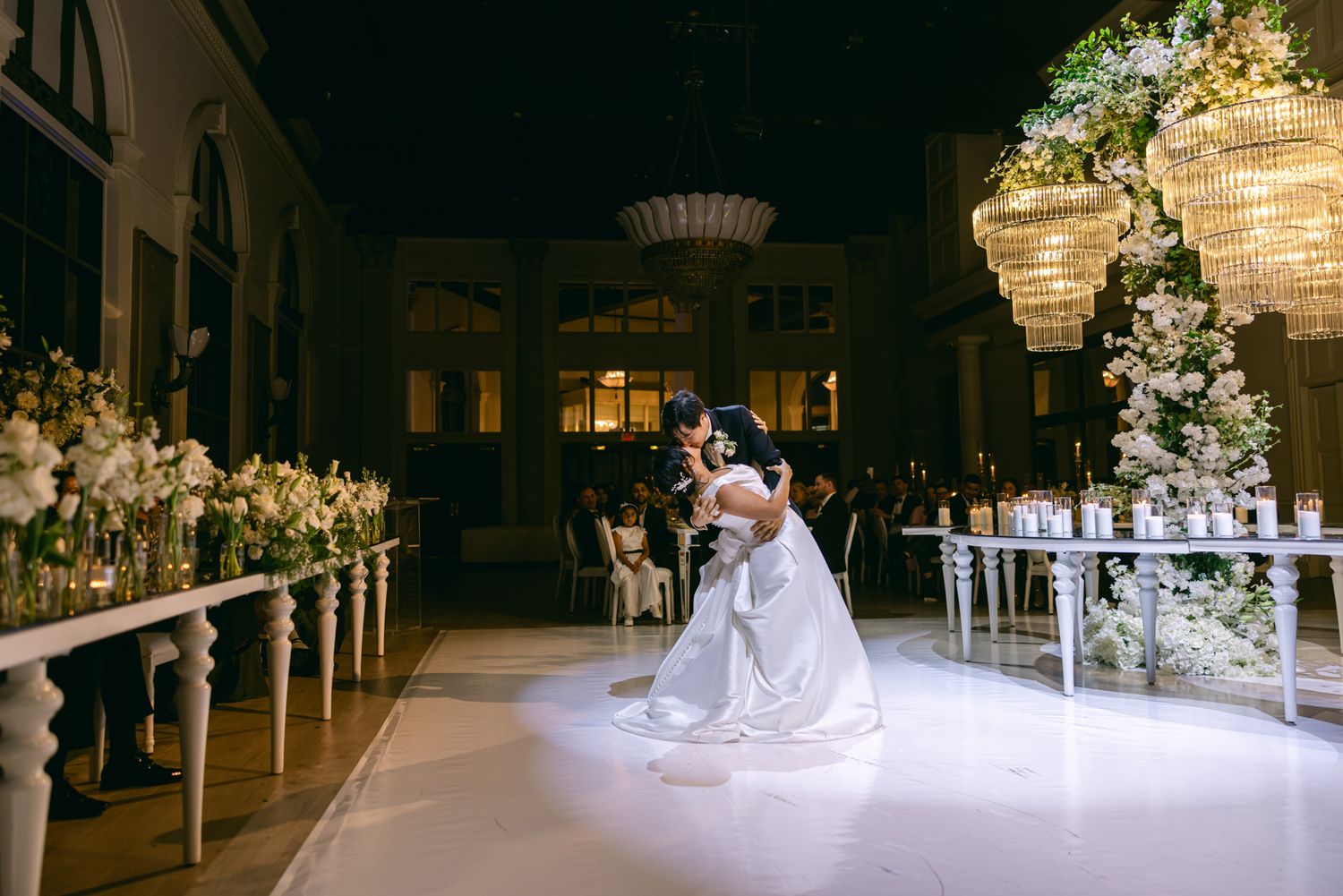 Bride and groom share a romantic first dance on a glossy ballroom floor surrounded by white flower arrangements and glowing candles.