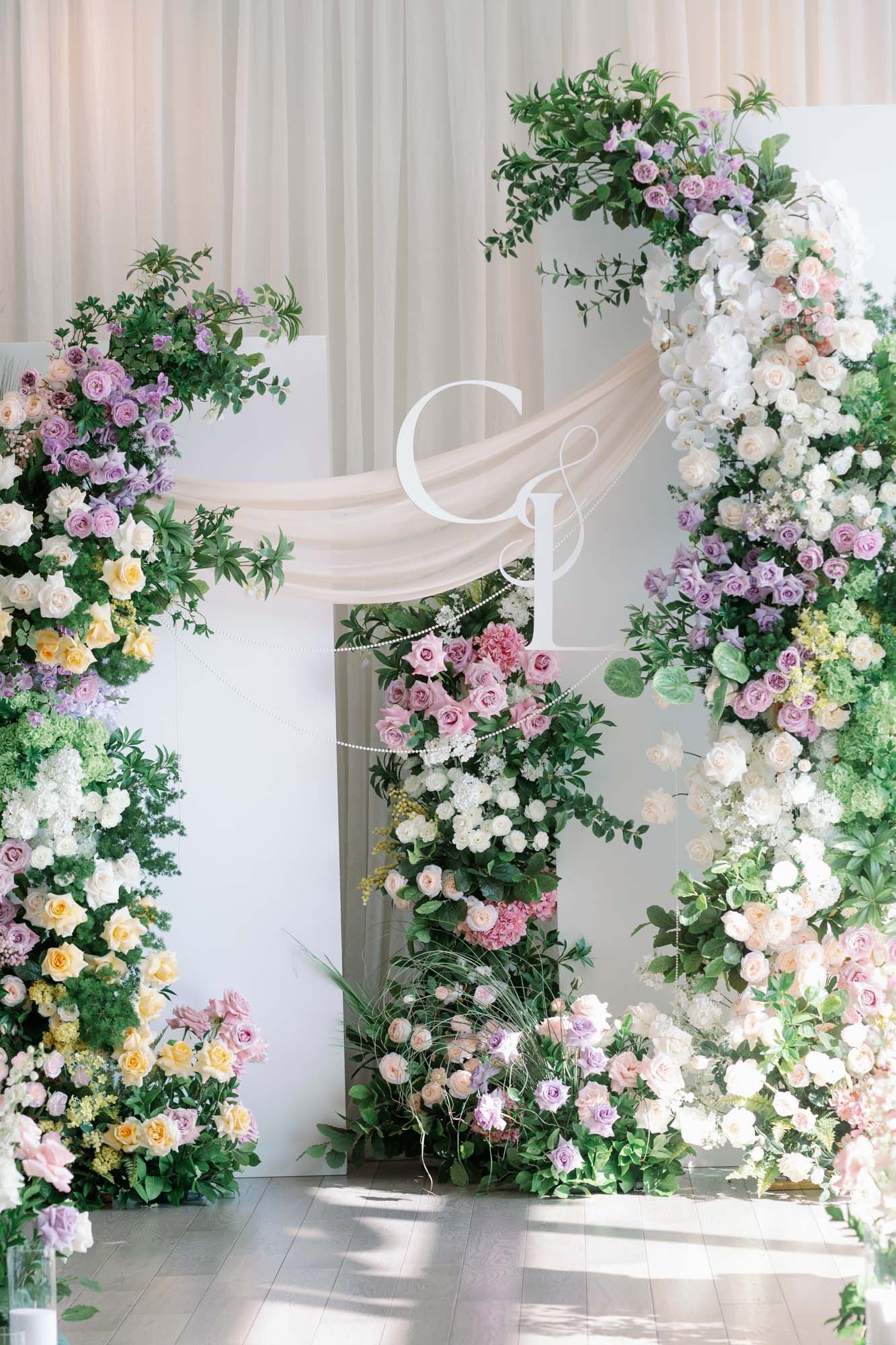 Elegant wedding arch decorated with lush purple, white, and yellow flowers with monogram signage.