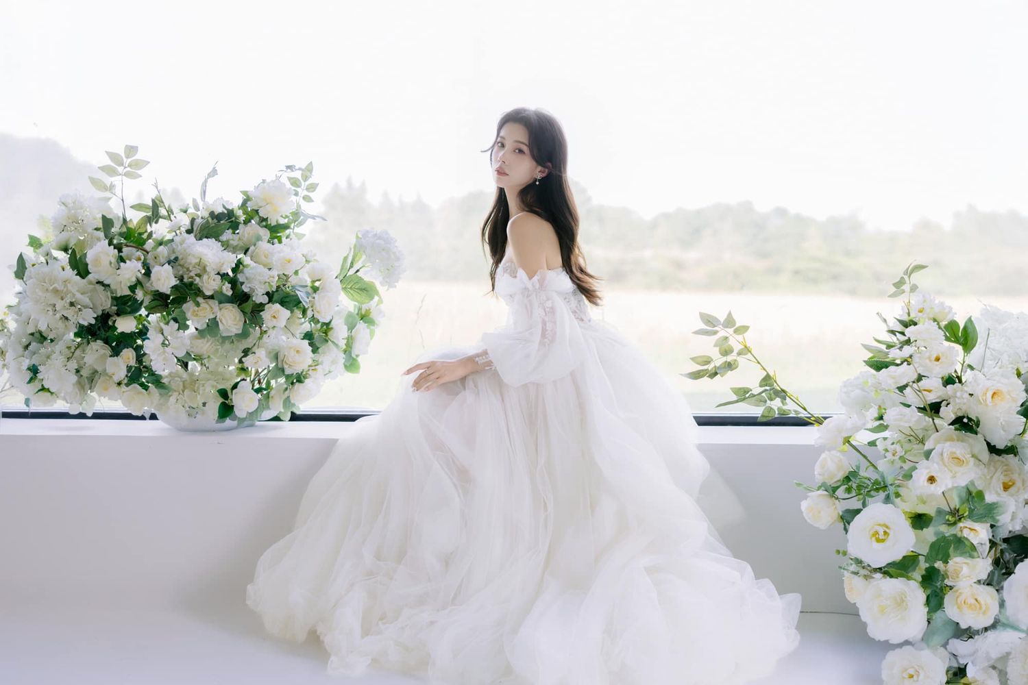 Elegant bride in flowing white gown surrounded by white floral arrangements in bright setting.