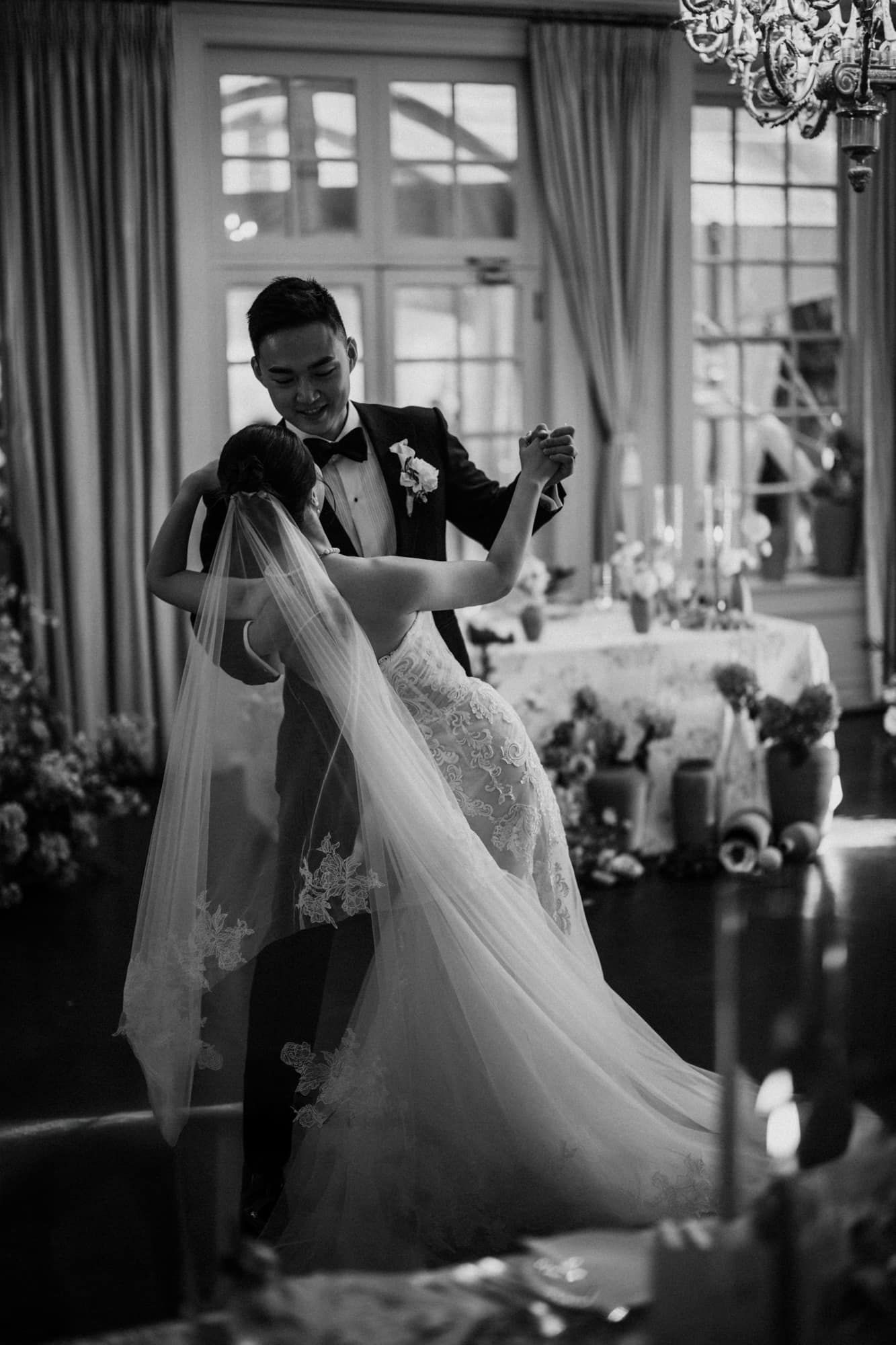 Black and white photo of groom spinning bride during their elegant first dance in a candlelit venue.