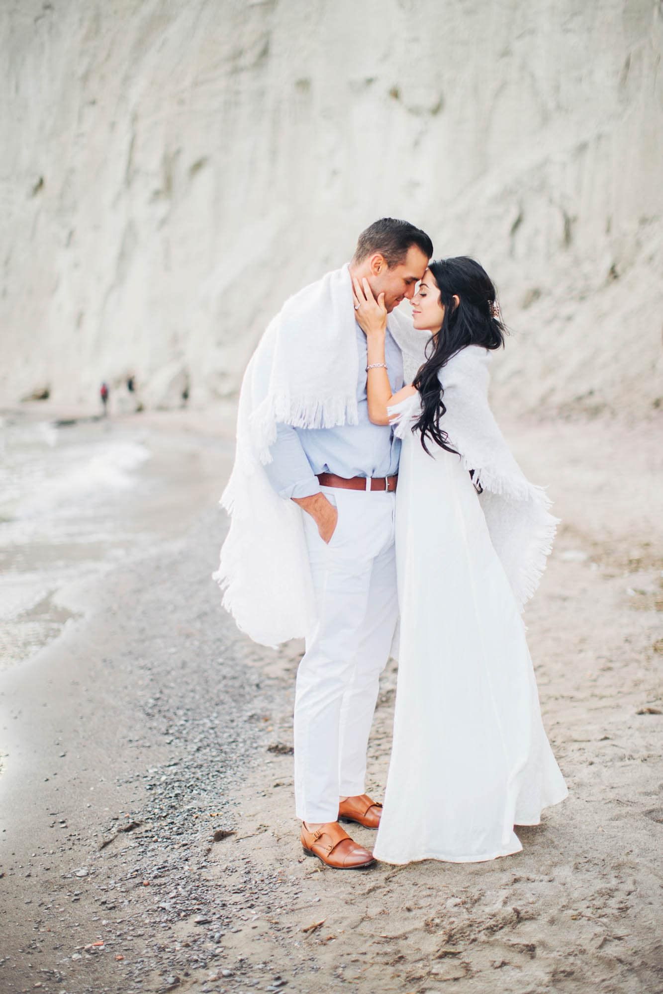 Couple shares a tender moment on a serene white chalk beach in flowing white attire.