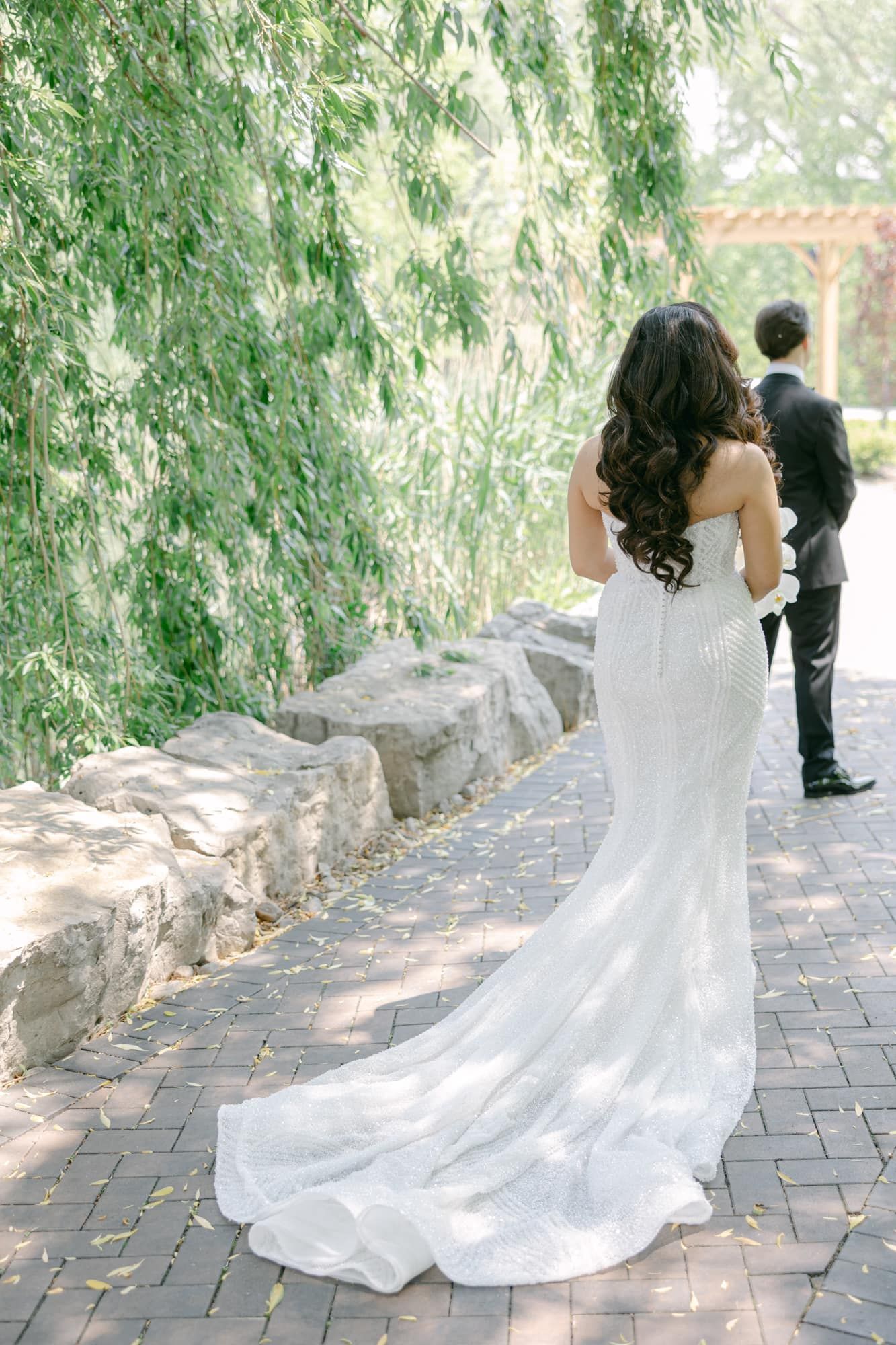Bride in strapless beaded mermaid gown with long train walks with groom along ivy-covered stone pathway.