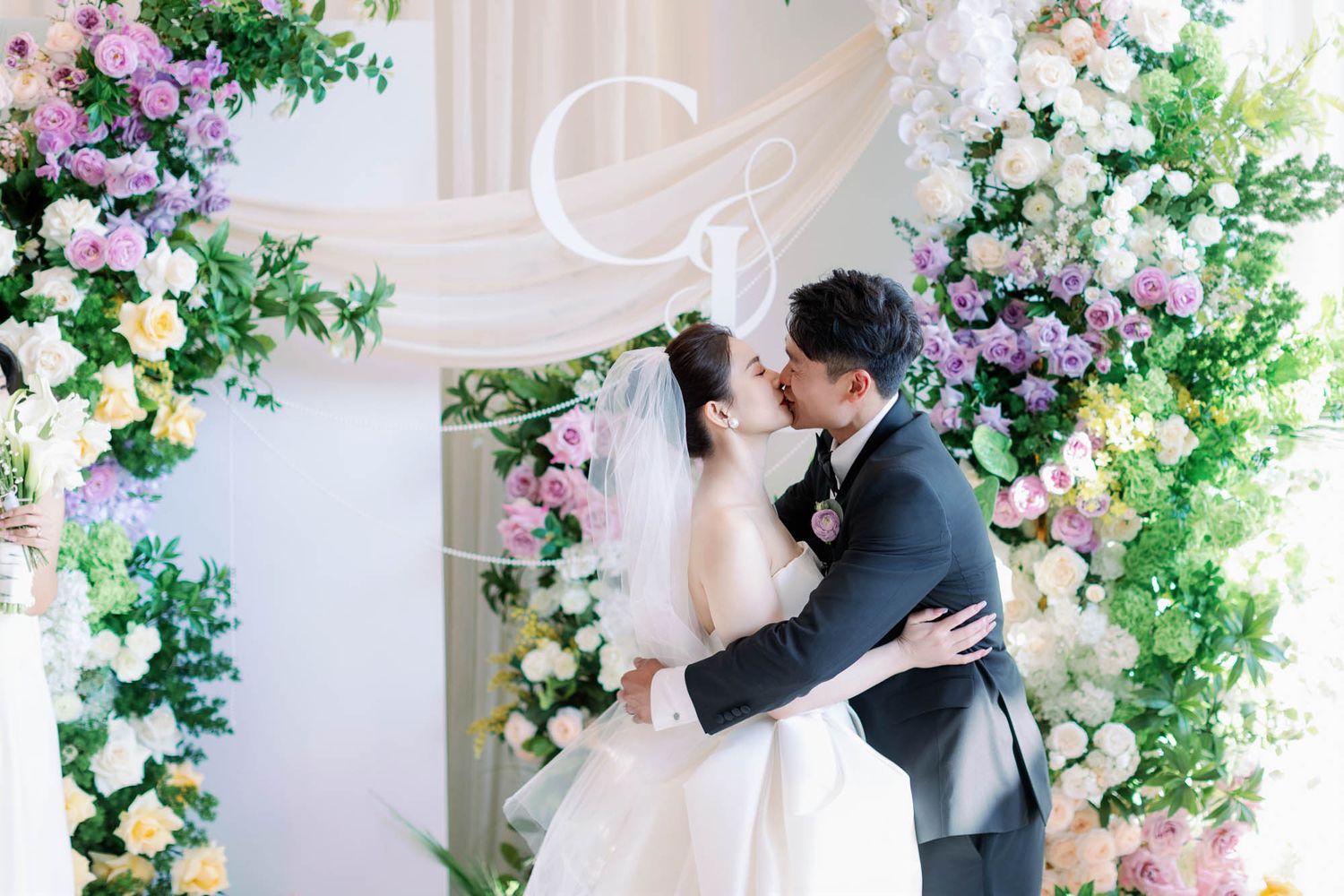 Bride and groom share first kiss under floral arch with colorful spring flowers at ceremony.