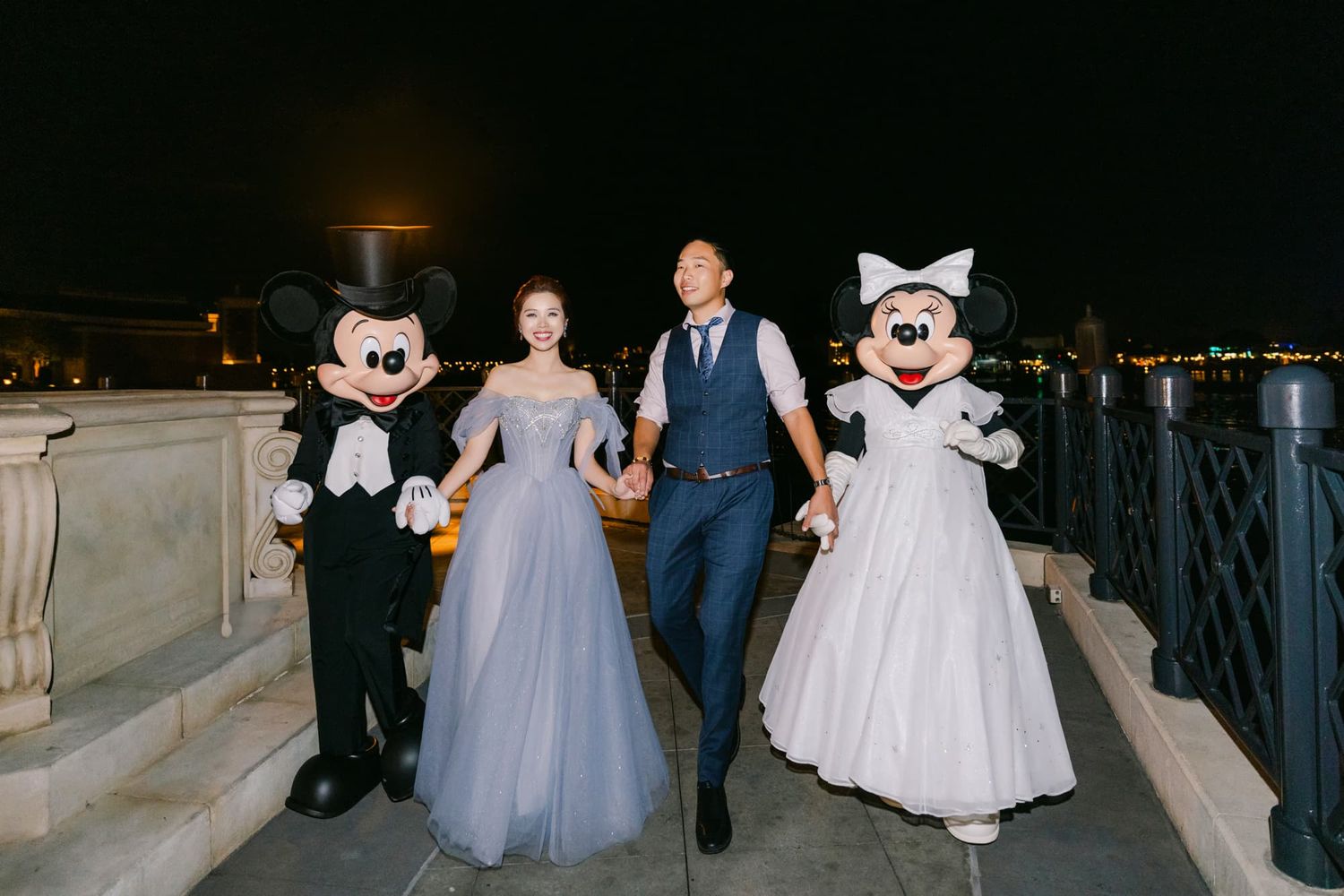 Couple in formal wear poses with Mickey and Minnie Mouse characters at night during a Disney wedding celebration.