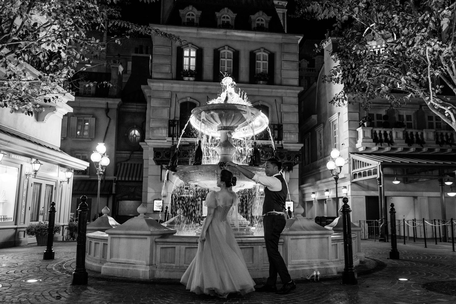 Black and white photo of bride and groom dancing by a glowing fountain at night.