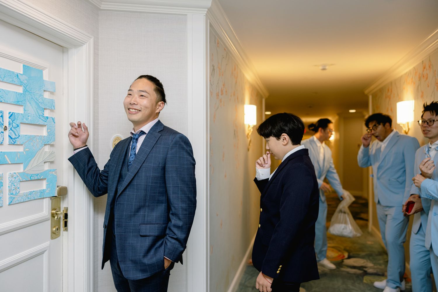 Groom in navy suit greets guests at door decorated with red double happiness symbol.