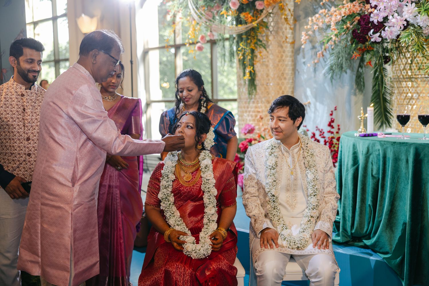 A bride in a red sari wearing a flower garland receives a blessing from an elder beside her groom during a multicultural wedding ceremony.