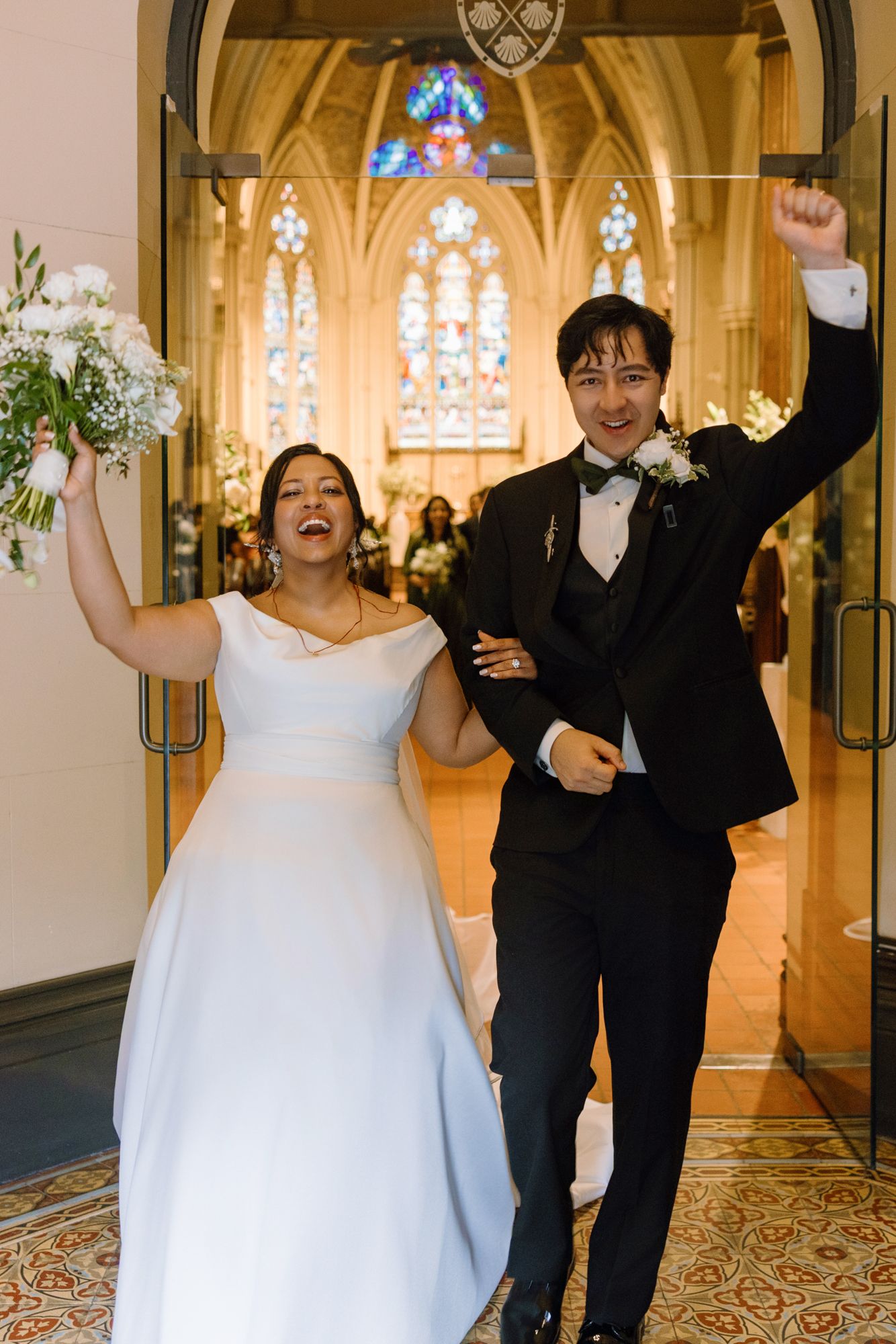 Smiling bride in a white gown and groom in a black tuxedo walk arm in arm out of the church, cheering with raised fists.