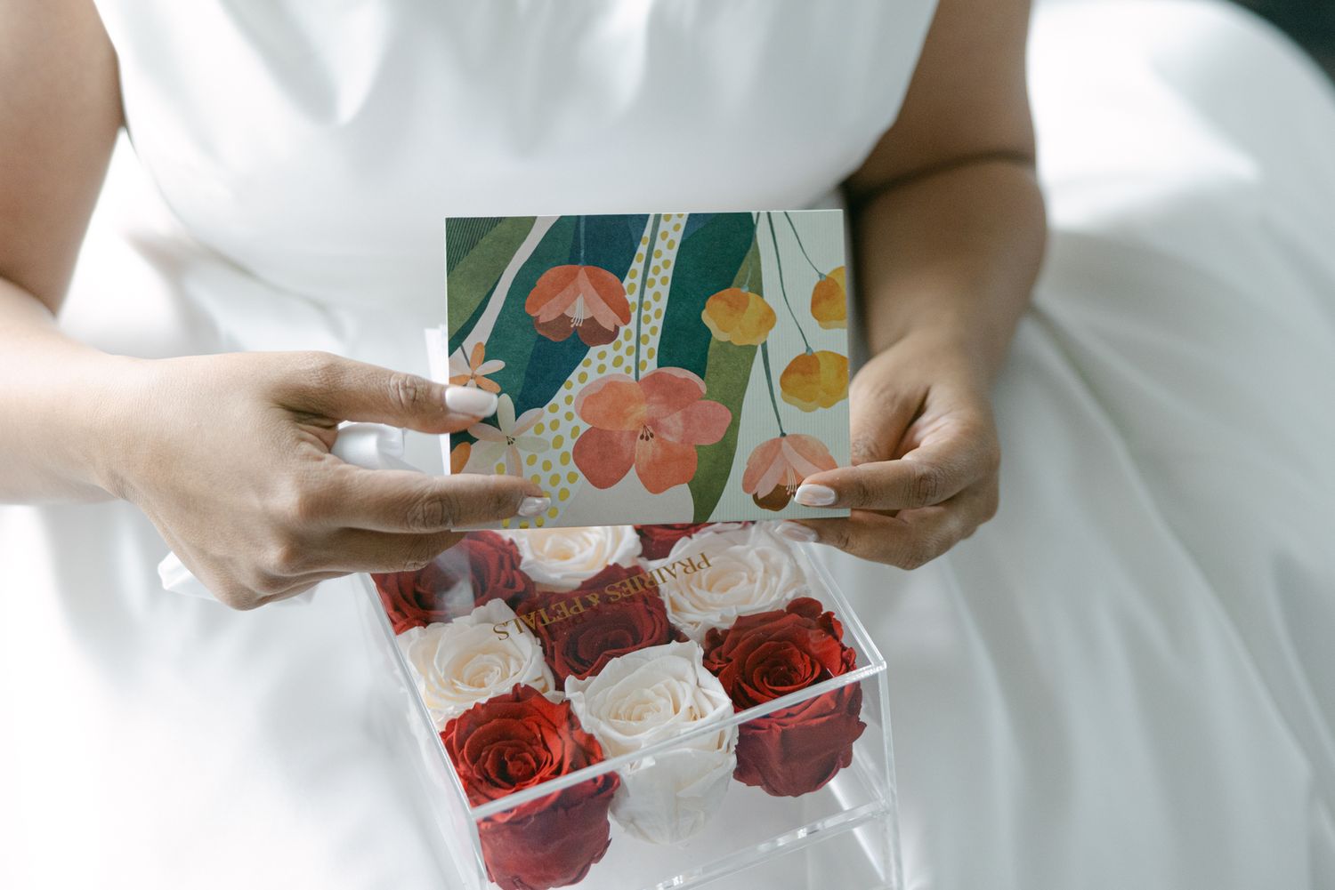 Bride in a white dress holds a clear acrylic box of red and white preserved roses with a floral-patterned lid.