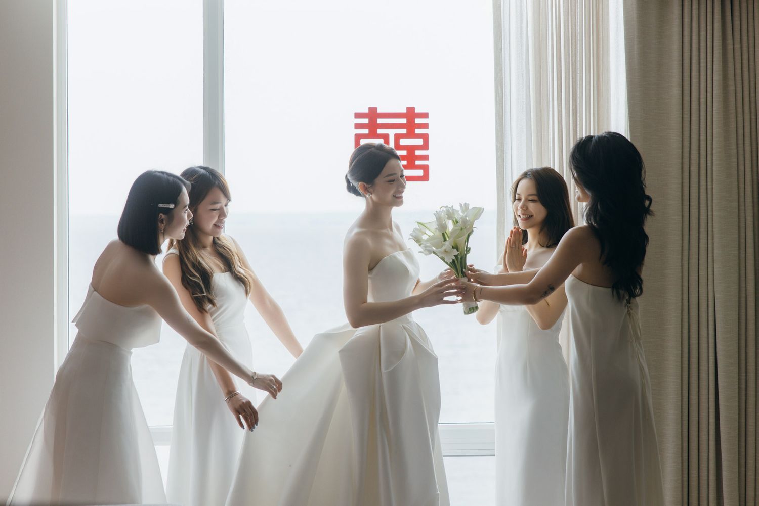 Bride in white gown with bridesmaids holding tulips near window with double happiness symbol.