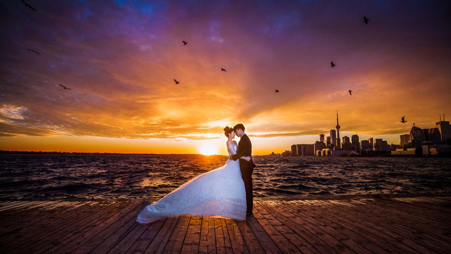 Couple embracing in wedding attire at sunset by the waterfront with a city skyline and birds in the colorful sky.