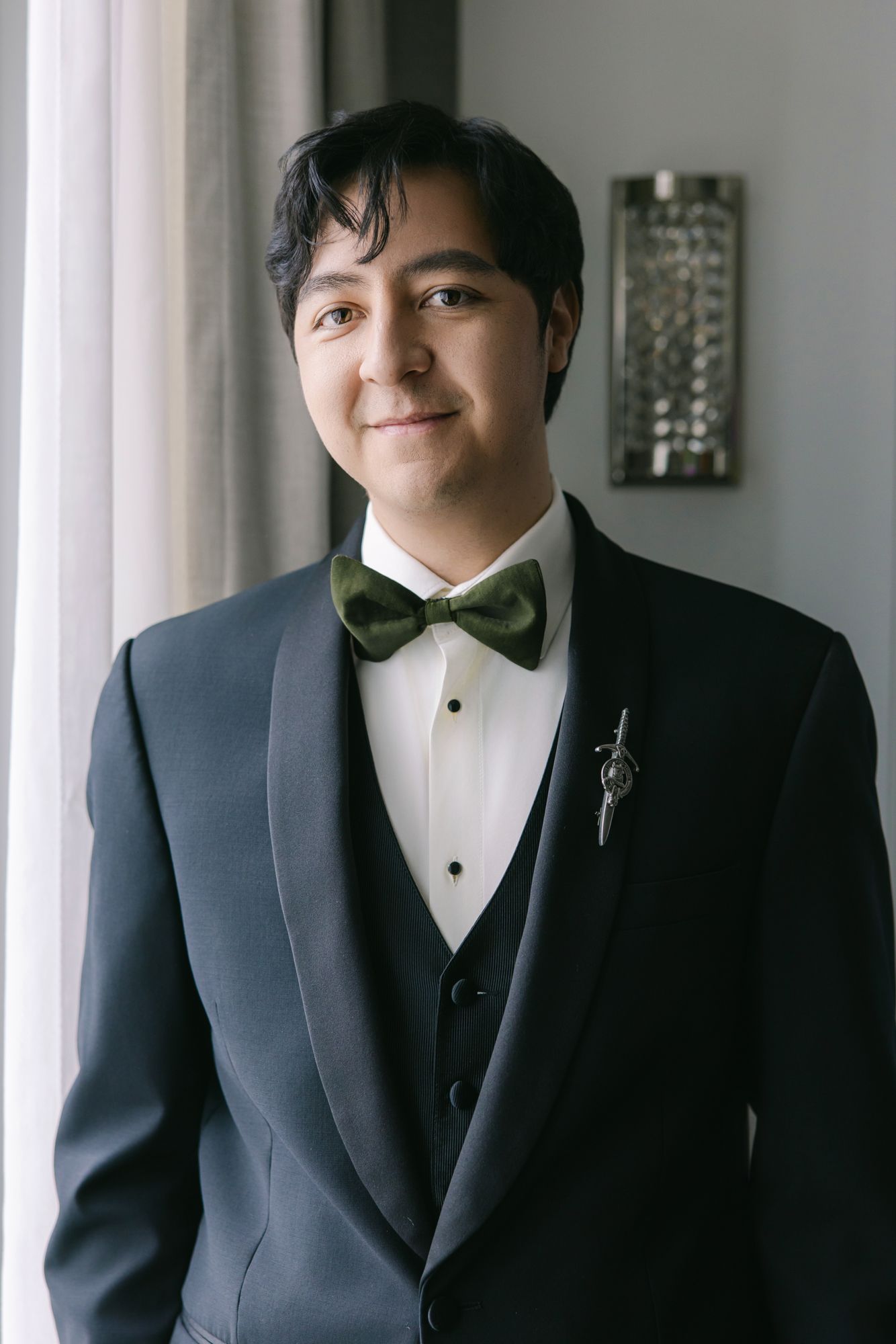 Groom in a dark shawl-collar tuxedo, green bow tie and silver boutonniere smiles beside a sunlit window before the wedding.