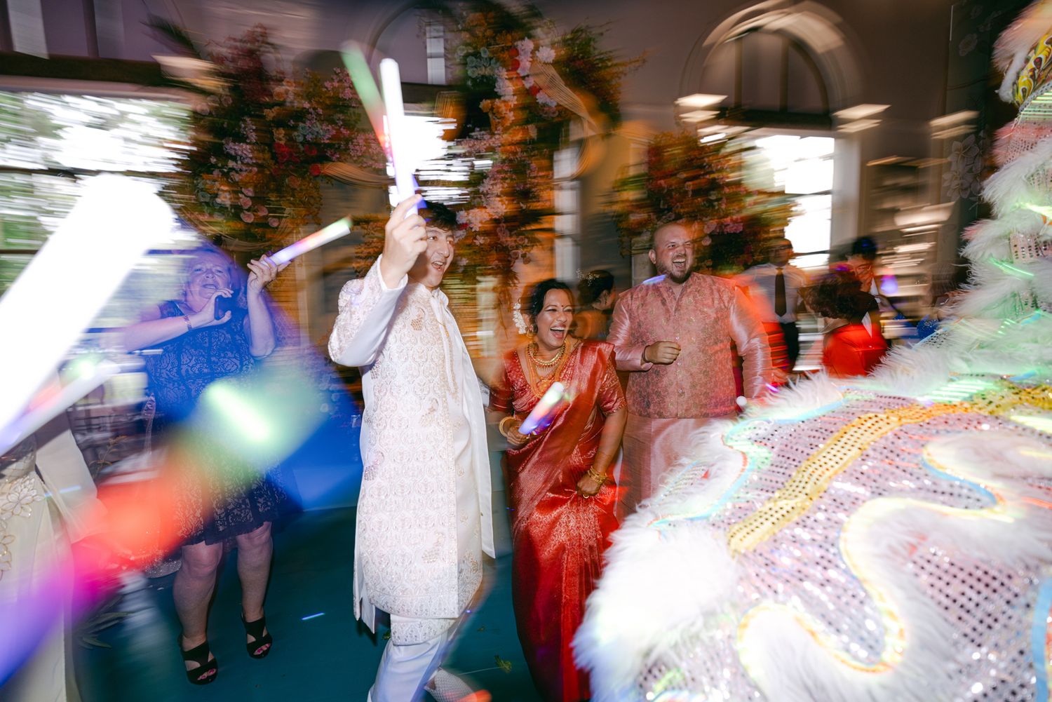 A lively wedding procession blurs with motion as a groom in white dances with guests and a lion dance costume nearby.