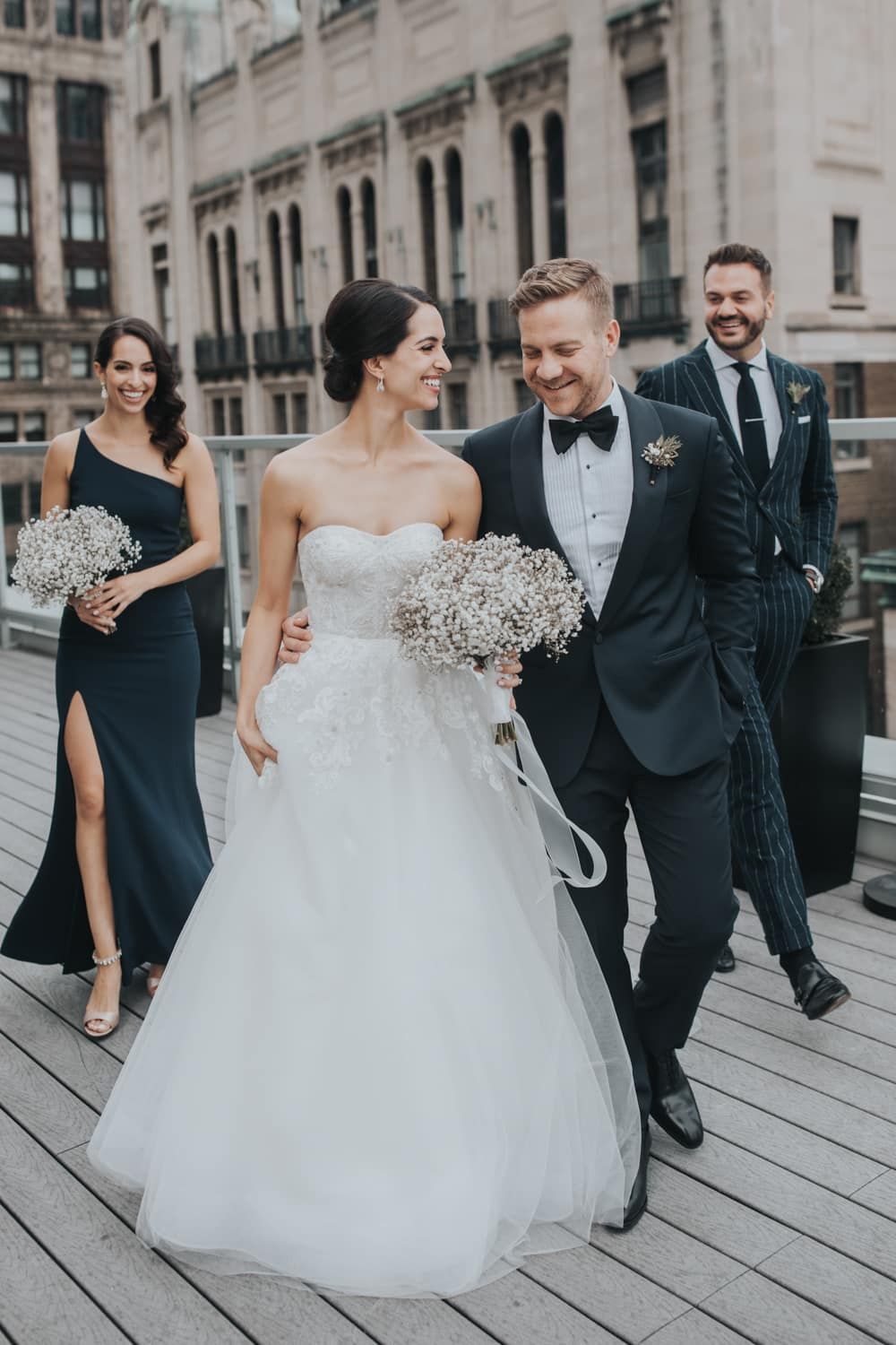 Smiling bride and groom walk with wedding party on wooden boardwalk in urban setting with bouquet.