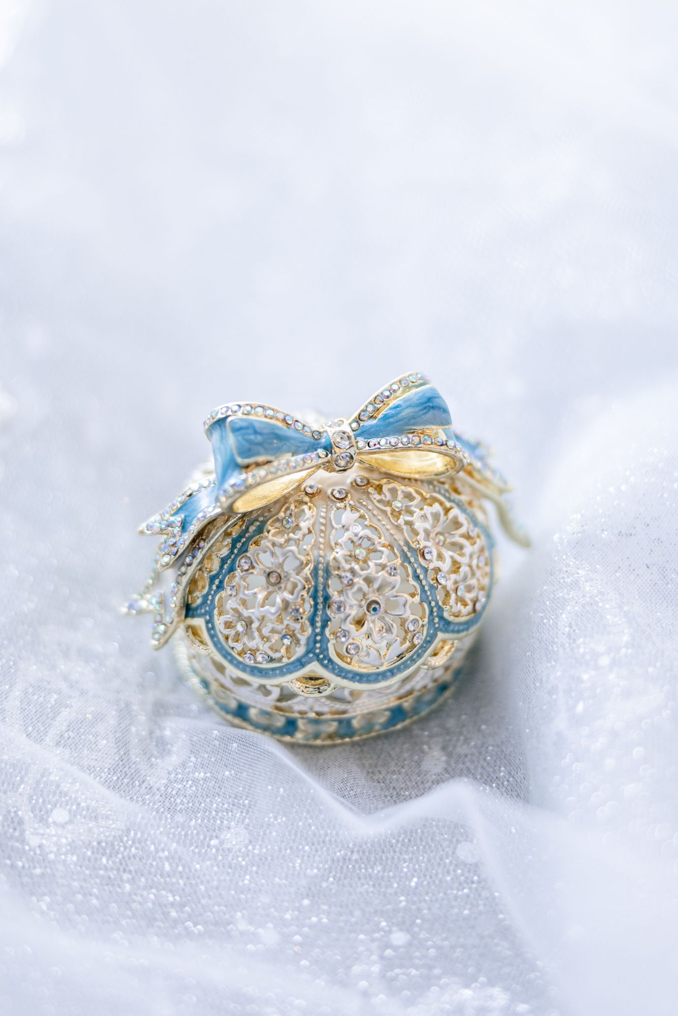 Elegant blue and gold Christmas ornament with intricate lace patterns and ribbon bow on white fabric background.