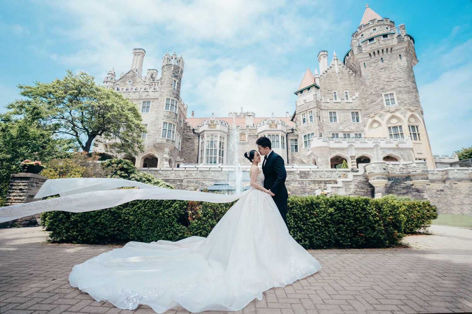 Bride and groom sharing a romantic kiss in front of a grand castle with manicured hedges outdoors.