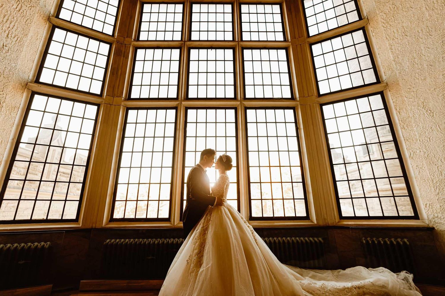 Bride and groom silhouetted against stunning tall arched windows in romantic castle wedding portrait.
